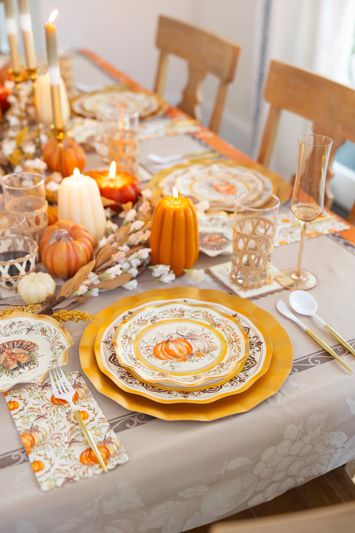 The Turkey & Gourds Table Setting by Sophistiplate features designer plates, gold chargers, candles, decorative pumpkins, glassware, and cutlery on a floral tablecloth with a festive fall centerpiece.